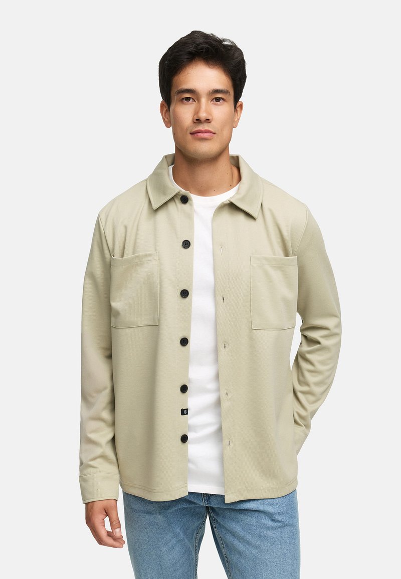 Lys grønn skjorte med krage, to brystlommer og svarte knapper, paret med en hvit T-skjorte og lyse denimjeans.