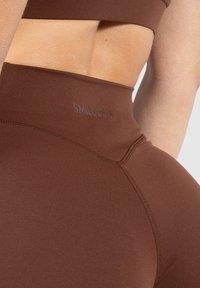 Bruine naadloze legging met een hoge taille, met een soepele textuur en een subtiele logo-accent op de tailleband. Close-upweergave benadrukt de stikseldetails.
