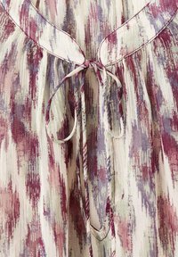 Blouse légère avec un motif floral dans des teintes de violet, rose et crème. Présente un col rond et un détail de nœud à l'avant.