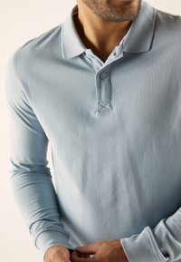 Svetle modrá dlouhá polo košile vyrobená z jemné tkaniny, s límečkem, tříknoflíkovým zapínáním a texturovaným povrchem.
