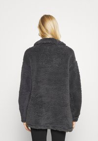 Chaqueta de forro polar gris oscuro con una superficie suave y texturizada, cuello ancho y corte holgado. Presenta hombros caídos y un dobladillo ligeramente curvado.