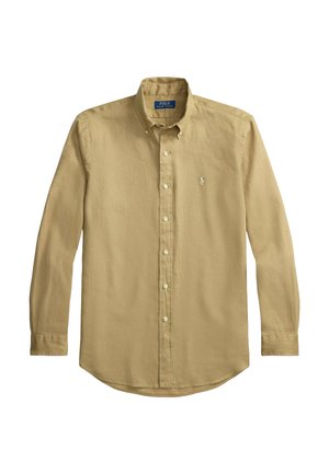 Man met krullend haar en bril draagt een mosterdkleurig overhemd, een groene trui over de schouders gedrapeerd, blauwe spijkerbroek, een bruine riem, horloge en bruine schoenen.