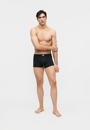 Vyriškis be marškinėlių stovi basas, dėvi juodos spalvos Adidas boxer šortus, nuleidęs kairę ranką ir išreikšdamas neutralų veidą priešais paprastą baltą foną.