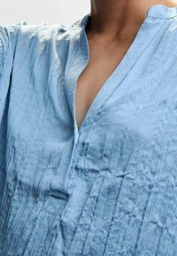 Blusa azzurra chiara realizzata in tessuto testurizzato, con scollatura senza colletto e dettaglio a bottoni sul pattino.