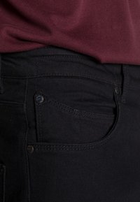 Jeans de mezclilla negra con una textura stone-washed, que cuentan con un bolsillo frontal y herrajes metálicos. La cinturilla muestra un ligero estiramiento y costuras detalladas.