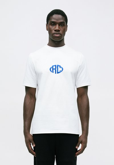 Mężczyzna stoi w pozycji frontalnej, ubrany w biały t-shirt z niebieskim logo "RC" umieszczonym na środku klatki piersiowej oraz w czarne spodnie, na tle jednolitego białego tła.