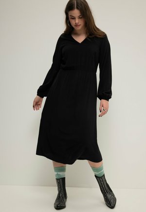 Abito lungo nero a maniche lunghe con scollatura a V e vita cinturata, che presenta un design fluido. Indossato con calzettoni verdi a motivi al ginocchio e stivali neri con borchie.