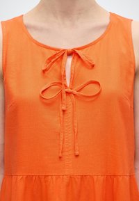 Ærmeløs orange kjole med rund halsudskæring, med to bundne sløjfer foran midt på og en rynket taljesøm.