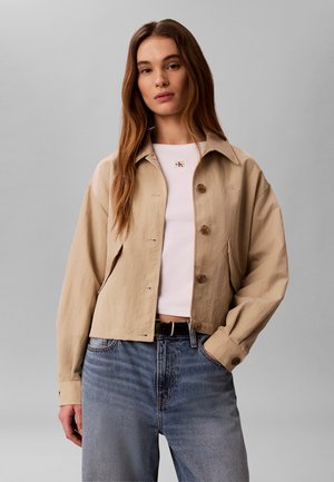 Frau trägt eine beige, geknöpfte Jacke über einem weißen, kurzen Oberteil und blauen Jeans, steht vor schlichtem grauem Hintergrund.