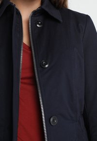 Námořnická modrá bunda s límcem, předním zipem a černými knoflíky. Materiál je hladký, s mírně strukturovaným střihem. Pod ním je červená košile.