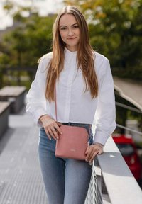 Růžová kožená kabelka přes rameno s texturovaným povrchem, s minimalistickým designem a jemným logem, nošená spolu s modrými džínami a bílou košilí.