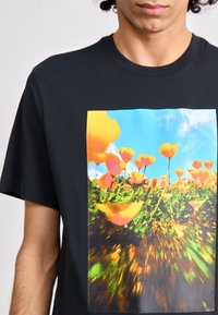 T-shirt preta com um gráfico vibrante de flores laranjas e cor-de-rosa contra um céu azul, com um efeito de movimento desfocado e folhagem verde.