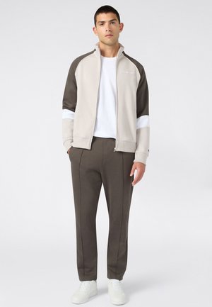 Modelo masculino vistiendo una chaqueta deportiva beige y marrón en bloque de colores, pantalones marrones, camiseta blanca y zapatillas blancas, de pie sobre un fondo liso.