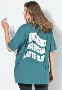 Tyrkysové oversized tričko vyrobené z bavlny, se silným bílým textem "ICED MATCHA LATTE CLUB" na zádech. Krátké rukávy a volný střih.