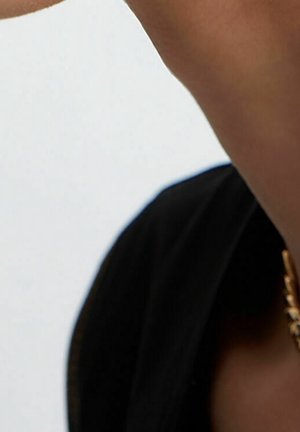 Femme vêtue de vêtements noirs et portant un collier chaîne en or sur un fond blanc, avec le bras levé près de son visage.