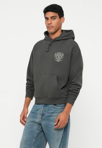 Férfi sötétszürke kapucnis pulóverben, amely elülső zsebes és a mellkasán embléma található, világoskék farmernadrággal párosítva, fehér háttér előtt áll.