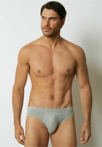 Grå bomullsbrief med en mjuk textur, med en sydd midja och en strukturerad passform. Modellen står med en neutral hållning.