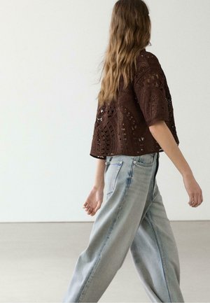 Donna che cammina, indossa un top corto lavorato a uncinetto marrone scuro e jeans larghi azzurri, con capelli lunghi ondulati e sfondo neutro.