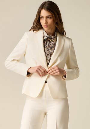 Kvinde der knepper en knap på en cremefarvet blazer over en mønstret bluse, iført matchende cremefarvede bukser, foran en neutral baggrund.