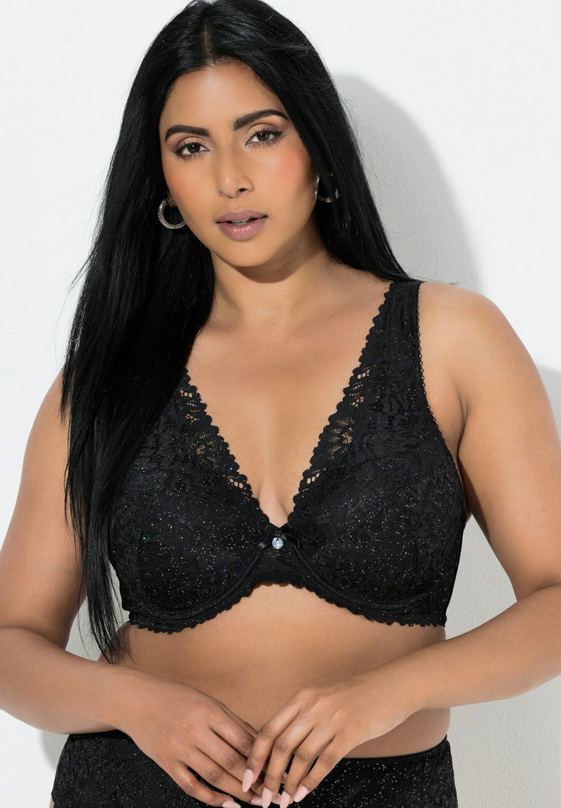 Kvinde med langt sort hår iført en sort blonde-bh og matchende undertøj, stående mod en hvid baggrund med hænderne foldet foran.