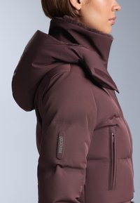 Bordeauxrode donsjas met een hoge kraag, voorzien van een zachte binnenkant en een zijvak met rits. RECCO®-logo op de linker mouw. Gladde stof.