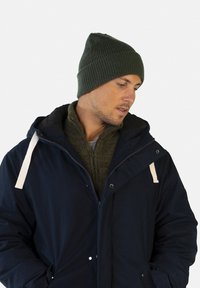 Un bonnet en tricot côtelé vert porté par un homme, associé à une veste à capuche bleu foncé et un pull zippé vert olive en dessous.