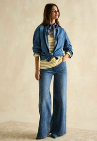 Jeanshemd, das an der Taille gebunden ist, über einem gelb-weiß gestreiften Hemd getragen. Weite blaue Jeans und ein blauer Bandana runden das Outfit ab.