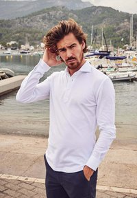 Witte langarm polo met knoopsluiting, gecombineerd met donkere broeken. Soepele textuur, casual pasvorm. De achtergrond toont boten en water.