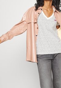 Ljust rosa, glansig vinylskjorta med långa ärmar och knappstängning, lagd över en vit V-ringad tröja med svarta prickar och grå jeans.