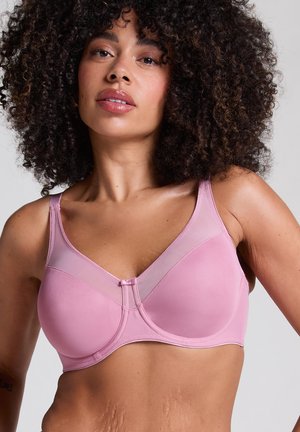 Vrouw met krullend haar, gekleed in een lichtroze beugel-bh met doorschijnende bovenpanelen, tegen een neutrale achtergrond.