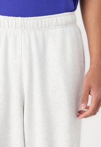 Pantalon de survêtement gris clair avec une taille élastique et des poches latérales. Le tissu est doux avec une texture subtile, comportant un petit détail de logo.