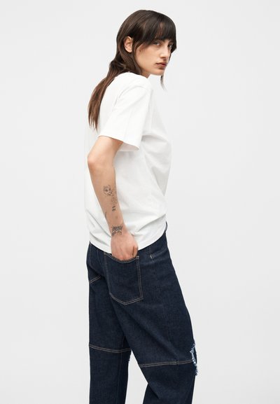 Άτομο με μακριά καστανά μαλλιά που φορά λευκό κοντομάνικο πουκάμισο και σκούρο φαρδύ τζιν με το χέρι στην πίσω τσέπη, δείχνοντας τατουάζ στο αριστερό χέρι.