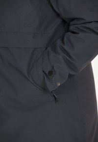 Mörk, strukturerad jacka med en dragkedjeficka och knappdekorer på ärmsluten. Tyget är slätt med en subtil lyster.