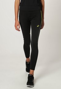 Černé sportovní legíny s žlutými akcenty, přiléhavý design a boční pruhy. Nosené s černými sportovními botami s logem.