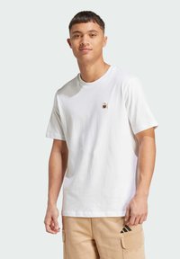 Hvid bomulds t-shirt med rund halsudskæring, der har et lille brunt bjørnesøm på brystet, korte ærmer og en afslappet pasform.