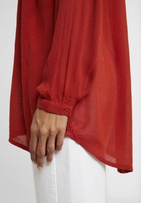 Main reposant à côté d’un pantalon blanc, portant un chemisier rouge transparent à manches longues avec poignets boutonnés et tissu texturé.