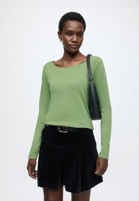 Zielona bluzka z długim rękawem i szerokim dekoltem, zestawiona z czarną spódnicą ze sztruksu z plisami oraz małą czarną torebką.