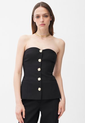 Kvinde iført sort stropløs top med fem gyldne knapper foran og matchende sorte bukser, accessoriseret med lagdelt guldhalskæde.