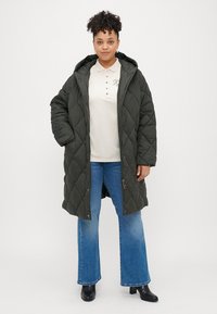 Duga podložena jakna u maslinasto zelenoj boji s dijamantnim kvadratnim uzorkom, koja ima kapuljaču, prednji zatvarač i velike bočne džepove. Nosi se preko svijetlog polo majice i širokih traperica.