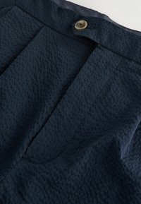 Détail de tissu texturé bleu marine avec une fermeture à bouton et des plis. Le matériau semble légèrement froissé avec une texture douce.