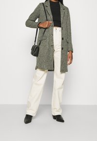 Abrigo gris de tejido con cuello muesca, botones frontales y bolsillos laterales, llevado sobre una blusa negra y pantalones anchos crema. Bolso negro y botas.