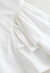Biela bavlnená látka so zdobením na rukáve v podobe nariasenej pufu, s jemnou textúrou a čistým prešitím.