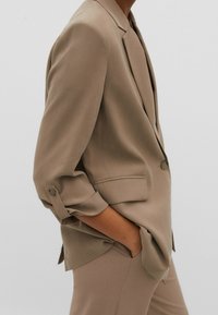 Blazer beige sur mesure avec un revers cranté, des manches retroussées et des poches avant. Tissu lisse et coupe décontractée mettent en valeur son design minimaliste.