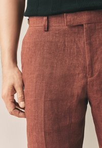 Pantalons en lin couleur rouille avec une texture lisse, présentant un devant plat, des poches latérales et une coupe ajustée.