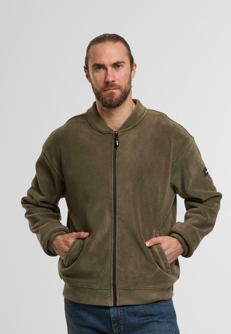 Oliwkowa polarowa kurtka z zamkiem z przodu, stojącym kołnierzem i bocznymi kieszeniami. Posiada teksturowane wykończenie oraz czarne akcenty metalowe.