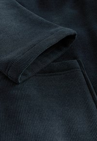 Tmavá ryhovaná tkanina s textúrovaným povrchom, zaobleným manžetom a detailom vrecka. Farba je hlboký, tlmený odtieň.