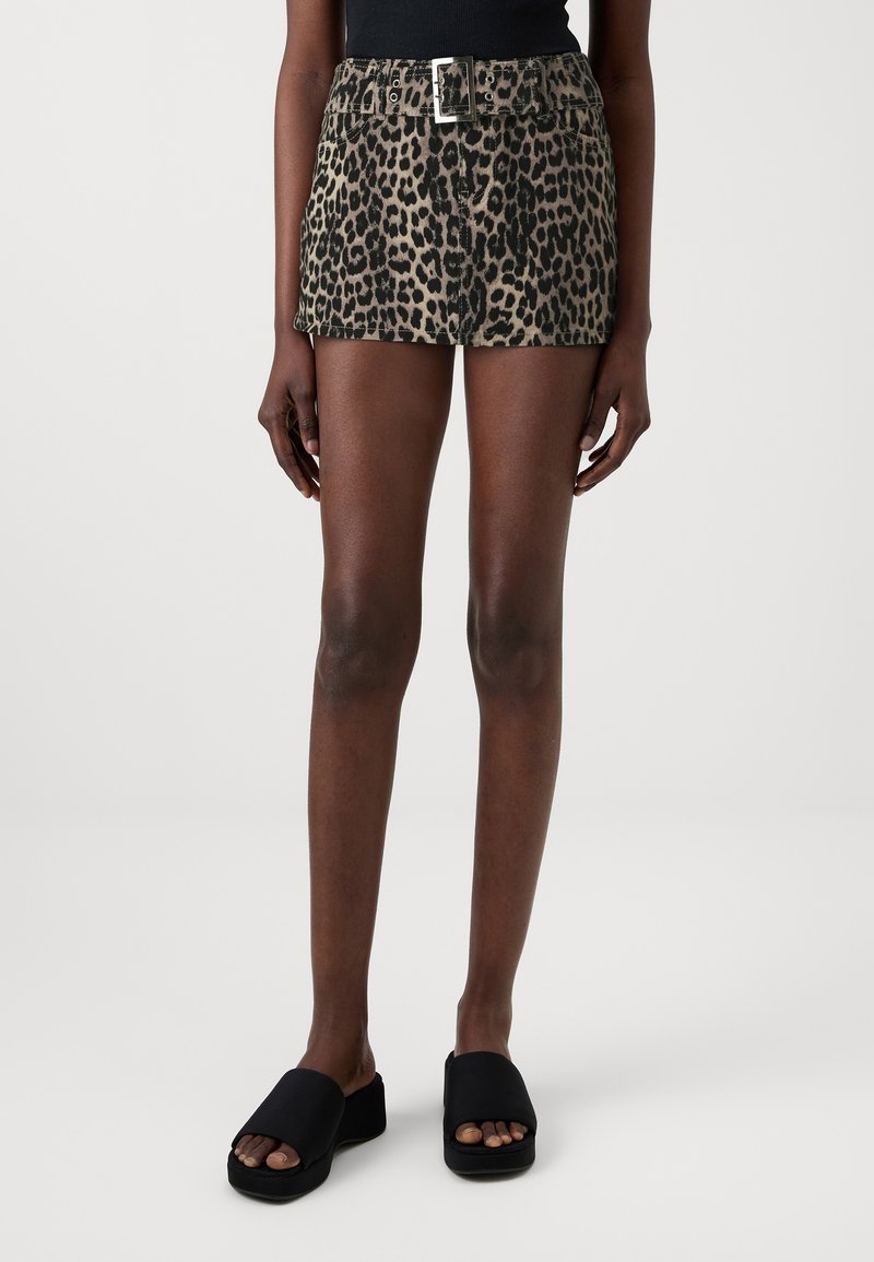 Femme portant une mini-jupe à imprimé léopard taille haute avec une ceinture, un top sans manches noir et des sandales noires à enfiler sur fond blanc.