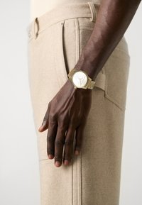 Reloj de pulsera dorado con una esfera circular, que presenta un diseño triangular. Se lleva encima de unos pantalones de lana beige con una textura suave y detalle de bolsillo lateral.