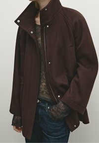 Persona che indossa una giacca zip marrone scuro sopra un maglione lavorato a maglia marrone semitrasparente e jeans blu scuro, con una mano in tasca.