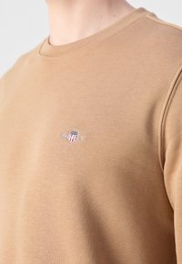 Gros plan d'une personne portant un sweat beige avec un petit logo écusson brodé et du texte sur la partie supérieure de la poitrine.